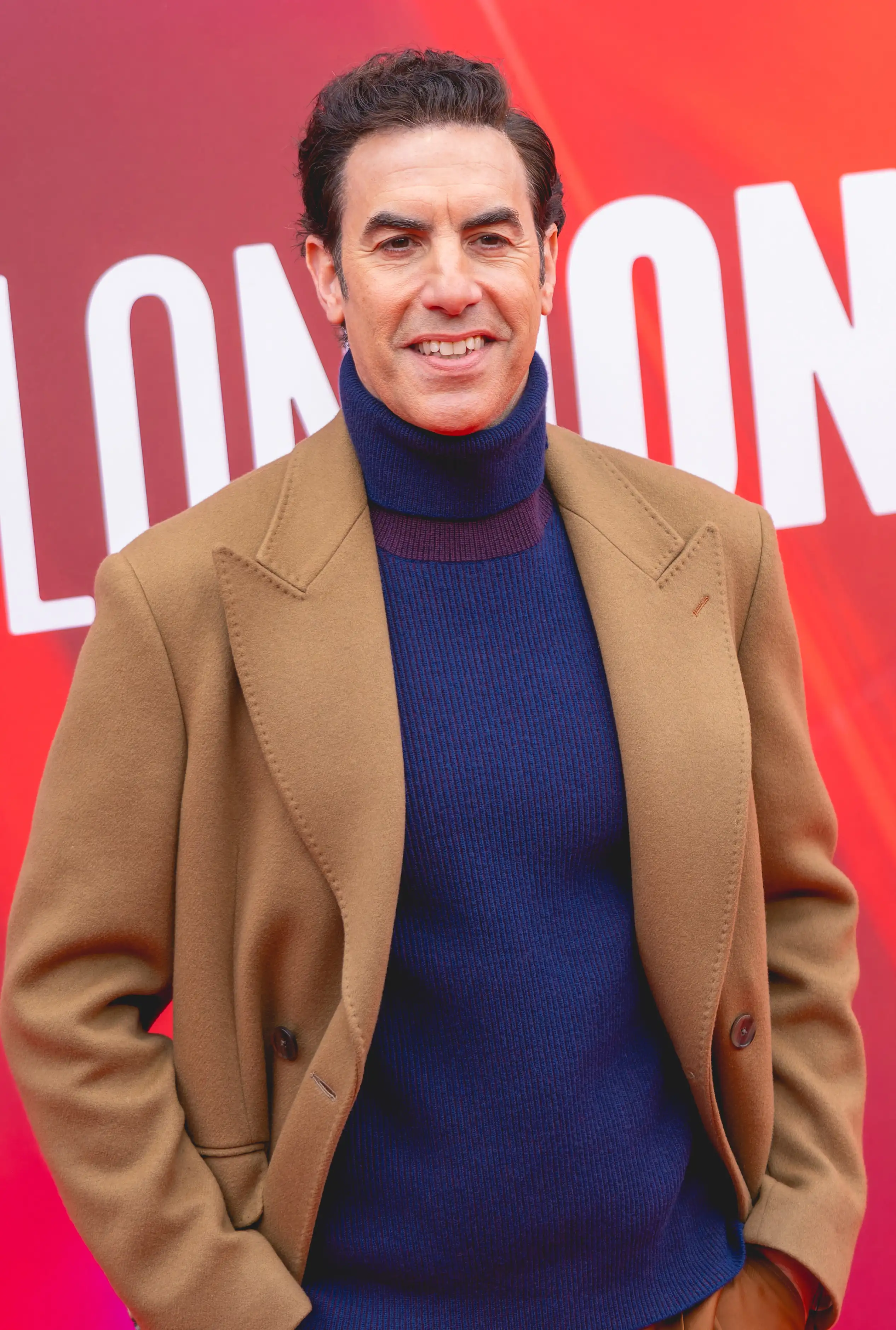 Sacha Baron Cohen in a camel coat and navy roll-neck, smiling, at the BFI London Film Festival, October 2024.
